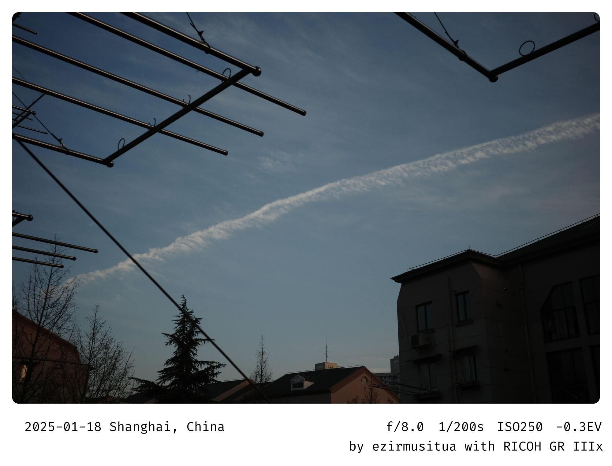 天空（sky）  飞机轨迹（contrail）  建筑（building）  电线（power lines）  树木（trees）  晴朗（clear）  宁静（serenity）  城市（city）  房屋（houses）  蓝天（blue sky）  人类活动（human activity）  对比（contrast）  现代（modern）  传统（traditional）  生活（lifestyle）  视野（view）